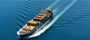 Cargo container ship sailing through calm blue ocean, stacks of colorful shipping containers on deck leaving a white wake behind.