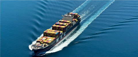 Cargo container ship sailing through calm blue ocean, stacks of colorful shipping containers on deck leaving a white wake behind.