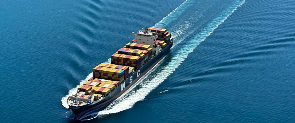Cargo container ship sailing through calm blue ocean, stacks of colorful shipping containers on deck leaving a white wake behind.